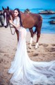 A woman in a wedding dress standing next to a horse on the beach.